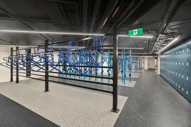 A modern commercial locker-room or staff changing facility with rows of blue metal garment racks and hooks, dark grey lockers, exposed ceiling services, linear LED lighting, and polished speckled flooring. The space has an industrial utilitarian fit-out and a clearly marked emergency exit.