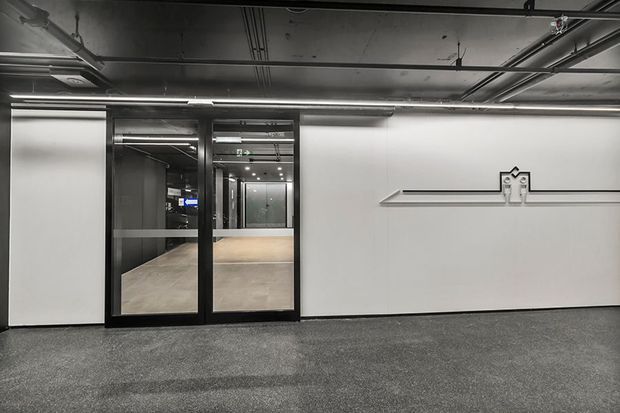 A contemporary commercial interior corridor or open office area with exposed services and a dark painted ceiling, grey carpet flooring, and a large white wall featuring a minimal linear graphic detail. Black-framed glazed double doors open into a brighter tiled space beyond. The space is predominantly monochrome, functional, and industrial in character.