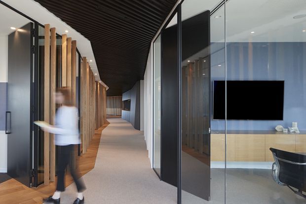 A contemporary interior corridor lined with vertical timber slat partitions and dark-framed glazed meeting or office rooms. The view includes a carpeted hallway, a dark linear ceiling feature, integrated timber cabinetry, and a wall-mounted television visible through glass. A blurred person is walking through the space.
