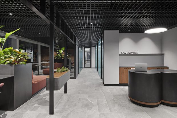 Contemporary commercial office reception and corridor with a dark exposed grid ceiling, full-height glazed meeting-room partitions, light grey stone-look flooring, integrated timber reception joinery, black curved reception desk, and built-in planter seating. The space has a refined, high-contrast commercial fit-out with layered lighting and greenery.