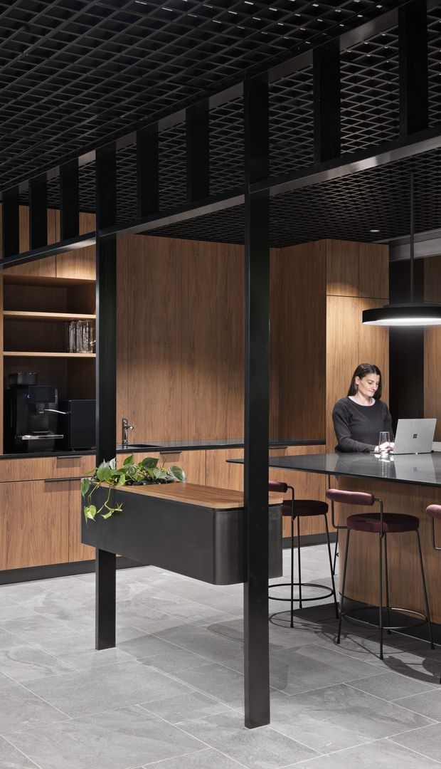 A dark, sophisticated commercial hospitality or office lounge interior with extensive walnut-toned timber wall panelling and cabinetry, black steel structural framing, and a black open-grid ceiling. A kitchenette or coffee station with shelving and coffee machine sits to the left, while a long stone-topped island or bar with burgundy upholstered stools occupies the right. A woman works at a laptop, and a black pendant light hangs above the counter. Large grey stone tiles cover the floor, with a planted built-in island adding greenery.