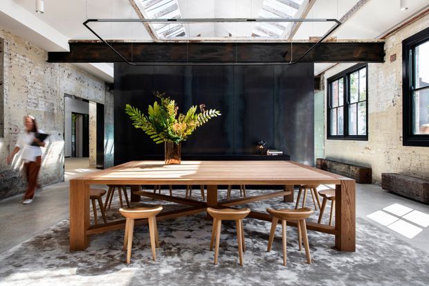 A bright, loft-like dining space centered on a large timber dining table with sculptural wooden chairs. Exposed pale brick walls, dark steel structural beams, a black feature wall, industrial-framed windows, and large roof skylights create a refined industrial interior. A floral arrangement provides a natural accent, while strong daylight casts geometric shadows across the concrete floor.