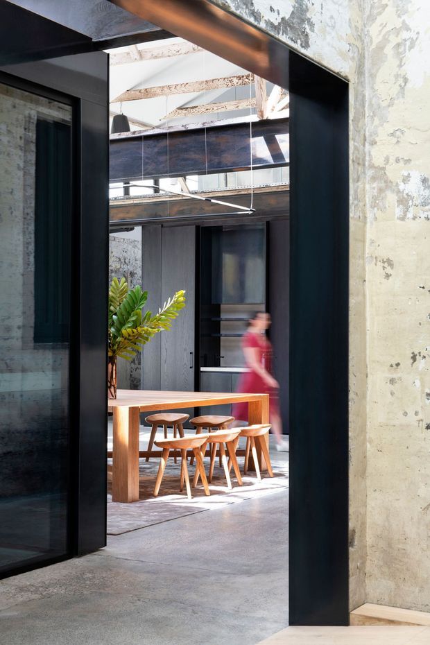 A view through a tall black-framed opening into an industrial-style dining area. The space features polished concrete flooring, rough exposed concrete walls, dark metal-framed glazing and doors, exposed structural beams, and a solid timber dining table with several timber chairs. A partially blurred person and indoor planting are visible in the background. Strong daylight enters through the high-level glazing and roof structure.
