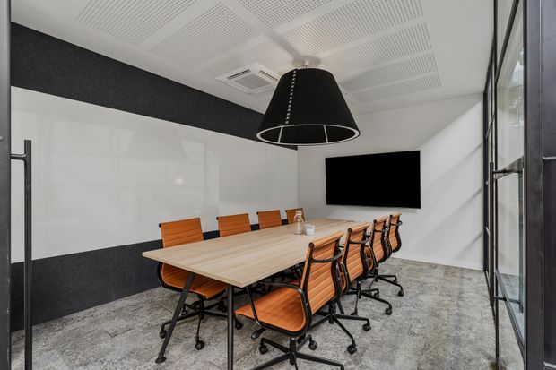 Contemporary conference room with a long light-wood meeting table surrounded by orange upholstered office chairs, a large black pendant light, wall-mounted display screen, acoustic wall panels, patterned acoustic ceiling tiles, polished concrete-style flooring, and full-height glazing.