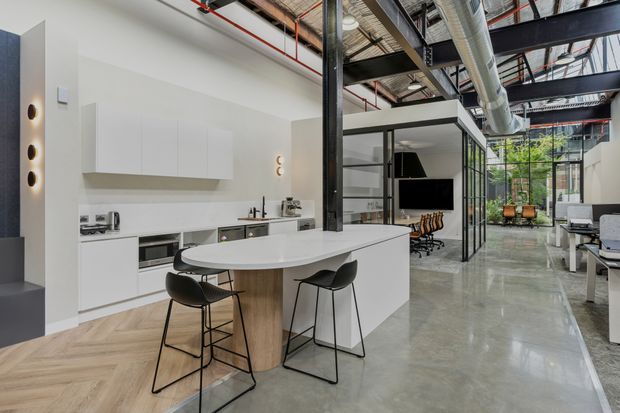 Open-plan contemporary industrial-style interior combining a white kitchen with a large island, casual bar seating, and an adjacent office or meeting area. Exposed black steel structural framing, visible services and ductwork, polished concrete flooring, extensive glazing, and a planted courtyard create a bright adaptive-workspace feel.