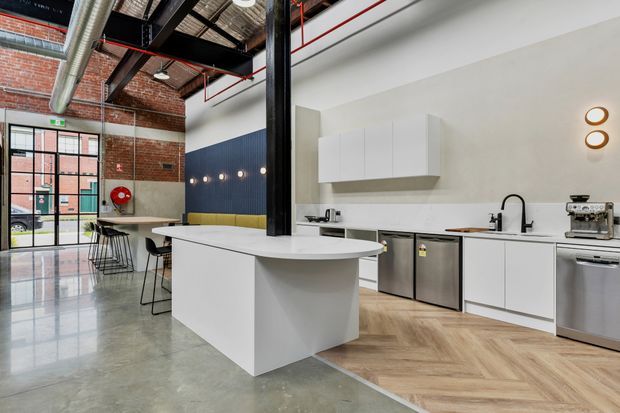 Open-plan industrial-style kitchen and dining area with a large white island, white flat-panel cabinetry, stainless-steel appliances, black tapware, and a herringbone timber floor zone. Exposed red brick walls, black steel structural framing, visible ductwork, mesh ceiling panels, and red service pipes create a warehouse-conversion character. Glazed industrial doors open to the exterior, while a navy upholstered banquette and dining table occupy the rear.