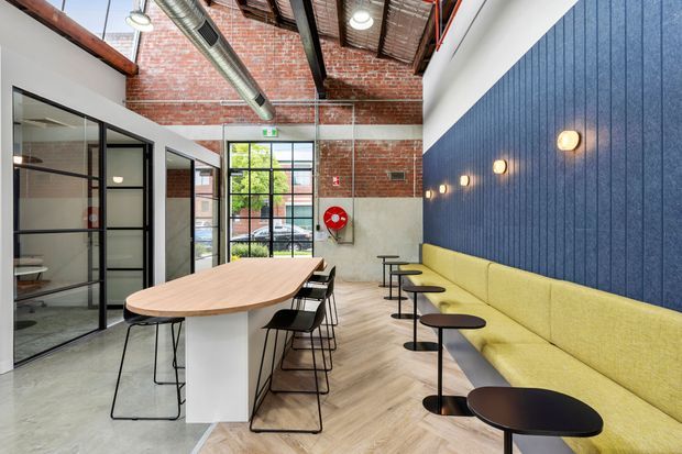 A bright industrial-style commercial breakout and dining area with exposed red brick walls, a high exposed timber and services ceiling, polished concrete and light timber flooring, black-framed glazed meeting-room partitions, and a long mustard-yellow upholstered banquette against a dark blue acoustic-panel wall. A large communal table with stools and small round café tables provide informal seating, while linear wall lights and exposed ductwork reinforce the workplace hospitality aesthetic.