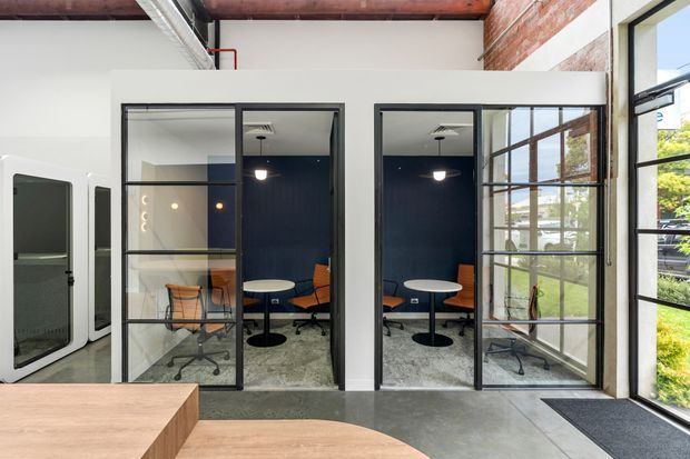 Contemporary office interior with two compact glass-enclosed meeting rooms, dark blue feature walls, black-framed glazing and doors, polished concrete flooring, exposed brick and timber ceiling elements, and views to greenery outside. A large timber work surface occupies the foreground.