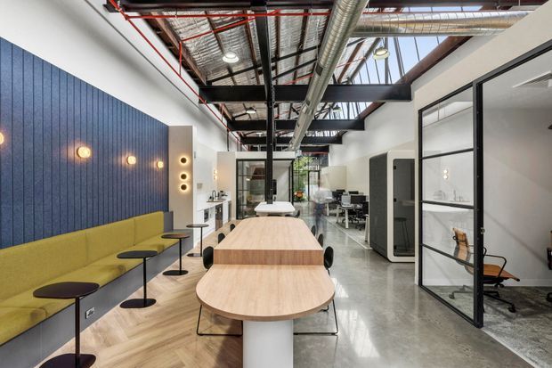 Contemporary commercial office interior with an open-plan collaborative dining and meeting area. The space features a long light-wood communal table, mustard upholstered banquette seating, dark blue vertically panelled feature wall, polished concrete flooring, glass-partitioned meeting rooms, exposed services and ductwork, black steel framing, and abundant glazing. The fit-out combines warm timber and upholstery with an industrial-style exposed ceiling.
