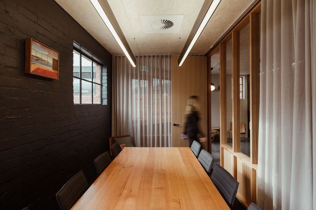 A narrow, refined dining room with a long light-wood dining table and dark upholstered chairs. The room features a dark painted brick feature wall, full-height sheer curtains, timber-framed glazed partitions, timber wall panelling, and linear recessed ceiling lighting. A doorway and glazed partition lead to an adjacent space.