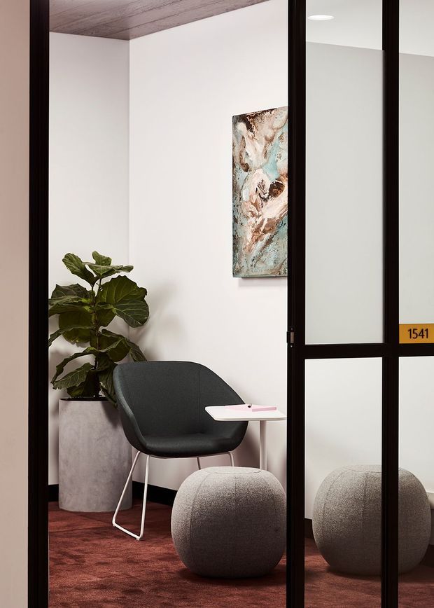 A compact, refined office or consultation nook viewed through a black-framed glazed partition. The space features a dark upholstered lounge chair, a small white side table, a rounded grey upholstered ottoman, a tall concrete-look planter with a leafy indoor plant, white walls, a timber-lined ceiling, and deep burgundy carpet. A framed abstract artwork and a yellow room-number plaque add small visual accents. The glazing reflects the seating and ottoman.