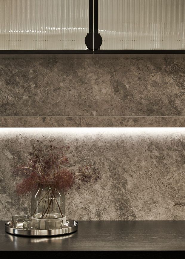 Close-up interior detail of a refined, moody feature wall clad in richly veined grey-brown stone or stone-look slabs. A concealed horizontal light strip washes the lower wall, while a dark countertop or shelf holds a metallic tray, glassware, and a clear vase with dried branches. Ribbed translucent glazing and a dark metal frame appear above.