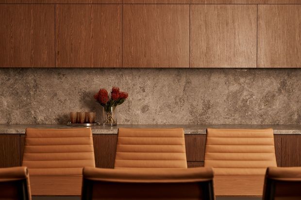 A warm, refined dining area with a long built-in wooden sideboard and upper cabinetry, a textured stone backsplash, and upholstered caramel leather dining chairs arranged around a table. Small floral and glassware accents sit on the counter.
