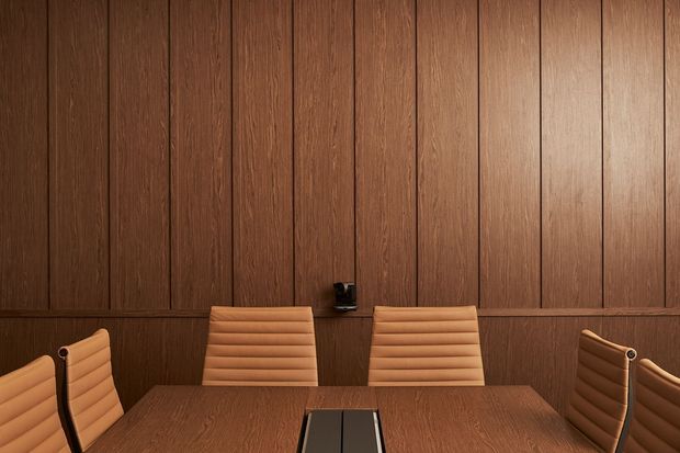 A warm, richly finished meeting or dining room with full-height dark timber wall panelling, matching timber table, and tan upholstered ribbed chairs arranged around the table. A central power or cable module is integrated into the tabletop, and a small wall-mounted fixture sits above it. The composition is refined and symmetrical with a subdued, professional atmosphere.