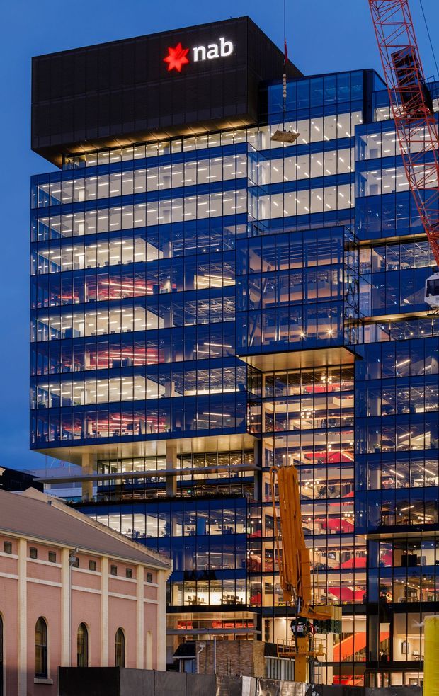 Twilight exterior view of a large contemporary commercial office building with extensive blue-tinted curtain-wall glazing, illuminated interior floors, projecting glass volumes, and a prominent black upper box displaying the NAB logo. A construction crane is visible alongside the building, with a preserved pink masonry structure in the foreground.
