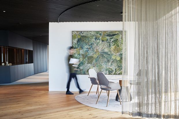 Contemporary interior dining area with a round table and upholstered chairs positioned beside full-height sheer curtains. A large abstract artwork forms a feature wall, while a dark slatted ceiling, timber flooring, and dark built-in joinery extend into the adjacent space. The image has a refined, high-contrast palette and abundant filtered daylight.