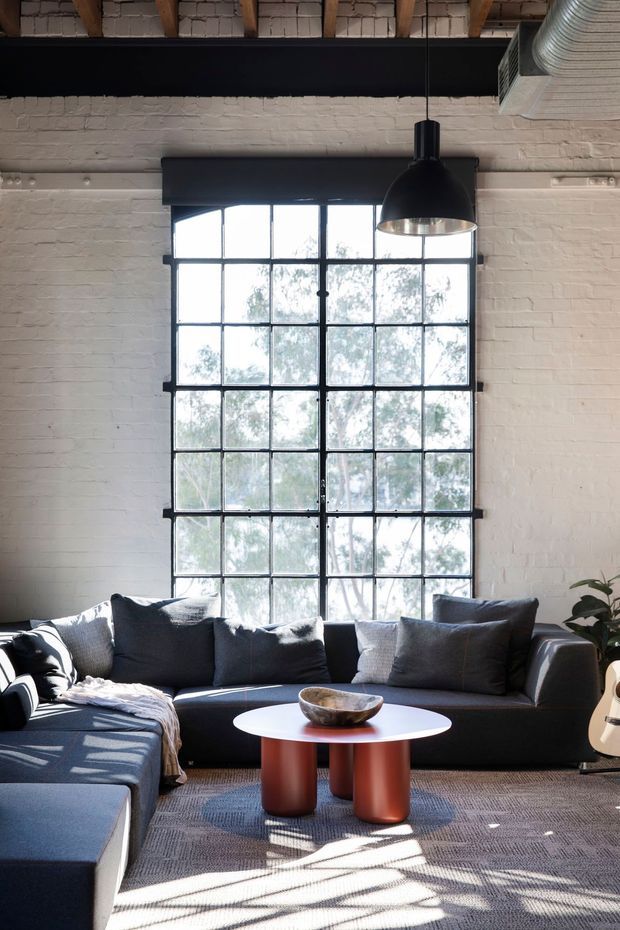 A refined industrial-style living room with a large black-framed multi-pane window, white-painted exposed brick walls, exposed timber roof structure, and visible ductwork. A dark charcoal sectional sofa surrounds a white round coffee table with cylindrical terracotta-toned legs. A large black pendant light hangs above, while strong daylight creates geometric shadows across the textured rug.