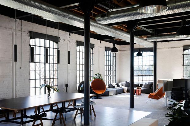 Open-plan industrial loft interior with white-painted brick walls, exposed timber roof structure, visible metal ductwork, black steel columns, tall multi-pane factory windows, and black pendant lighting. The space combines a large communal dining or work table with an informal lounge area and orange accent chairs.