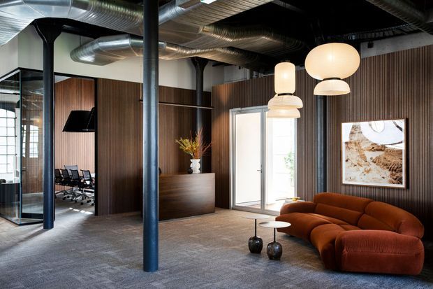 A sophisticated industrial-style commercial lounge or reception area with exposed silver ductwork, dark structural columns, charcoal ceiling, grey carpet, and extensive dark timber wall panelling. A sculptural rust-brown curved sofa and nesting coffee tables sit beneath layered cream pendant lights. Glazed meeting-room partitions, a timber reception counter, and framed artwork are visible.