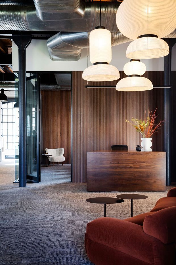 A sophisticated open-plan commercial reception or lounge area with exposed galvanized ventilation ducts, black steel framing, dark vertical timber wall panelling, and a large timber reception desk. Sculptural cream pendant lights hang overhead, while a deep rust-red sofa, low black coffee tables, and a sculptural armchair furnish the carpeted space. Glazed partitions reveal an adjacent seating area and industrial-style architectural details.