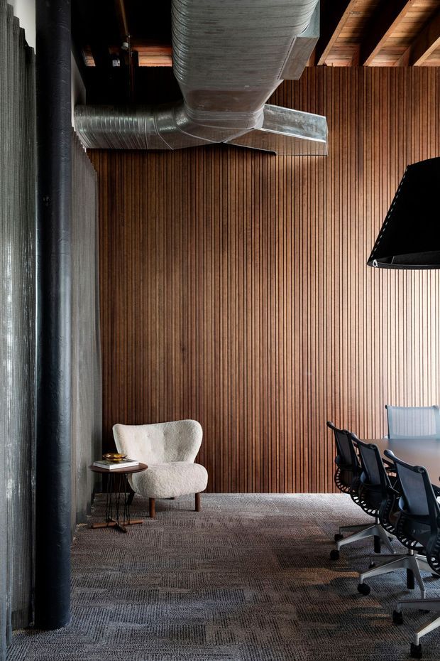A sophisticated industrial-style office or meeting room with exposed galvanized HVAC ductwork and black services against a warm vertical timber-slat feature wall. The space includes a long meeting table with black ergonomic office chairs, a sculptural upholstered lounge chair with a small side table, exposed timber roof framing, and dark textured carpet flooring. The contrast between warm timber, raw metal ducting, and black fixtures creates a refined yet utilitarian atmosphere.