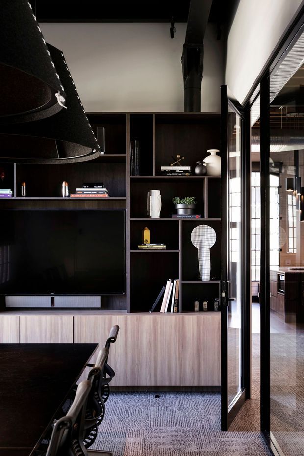 A refined contemporary living and dining area featuring a dark built-in entertainment and display wall, timber-look lower cabinetry, integrated television, open shelving, and sculptural decor. A large black pendant light hangs above the dining table, while black-framed glazed doors open toward a bright adjacent space. The room has a polished, moody palette with grey patterned carpet and strong vertical joinery lines.