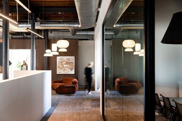 A sophisticated commercial interior with an open reception or lounge area, exposed timber ceiling structure, visible metal ductwork, dark structural columns, glass partitions, and warm brown carpet. A cluster of sculptural pendant lights illuminates rust-coloured lounge chairs, while a meeting room with office chairs is visible behind glazing. The palette is warm and refined with strong industrial architectural detailing.
