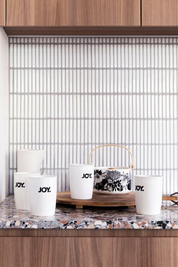 Close-up of a styled kitchen or kitchenette benchtop with white ceramic cups and a floral-patterned handled vessel arranged on a wooden serving board. The backdrop is a vertically ribbed white tile splashback, with warm timber cabinetry above and below and a multicoloured terrazzo-style countertop.