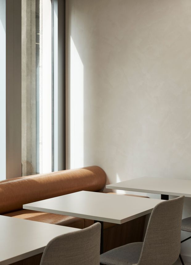 A minimalist contemporary dining or café-style seating area with a warm tan leather banquette beside tall glazing, paired with pale rectangular tables and upholstered grey chairs. The space has smooth warm off-white plaster walls, abundant natural light, and restrained neutral detailing.