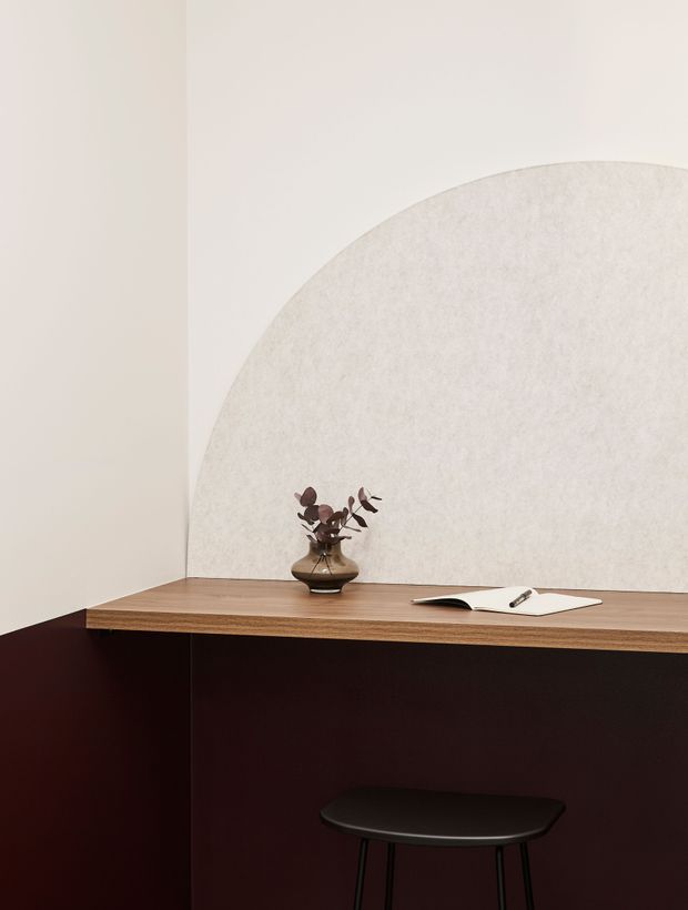 A compact, minimalist workspace with a wall-mounted or built-in timber desk spanning a corner. The lower cabinetry or desk base is deep burgundy, while the walls are warm off-white with a large textured semicircular feature panel. A small dark stool, ceramic vase with dried foliage, and open notebook complete the restrained composition.