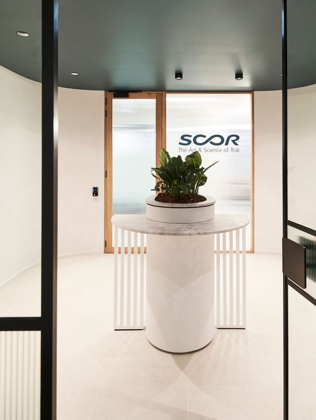 A refined commercial reception entry with a central cylindrical white reception table and planter, surrounded by curved white walls, pale flooring, dark charcoal ceiling, black-framed glazed partitions, and warm timber-framed frosted glass doors. The SCOR logo is visible on the rear glazing. Vertical white balustrade-like detailing and concealed/recessed lighting add a clean, contemporary corporate aesthetic.