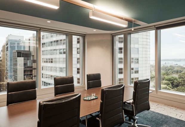 A contemporary high-rise meeting or dining room with a large rectangular wood table, black leather swivel chairs, expansive corner glazing, and city and waterfront views. The room features a dark teal ceiling, recessed and linear pendant lighting, neutral walls, and a dark area rug.