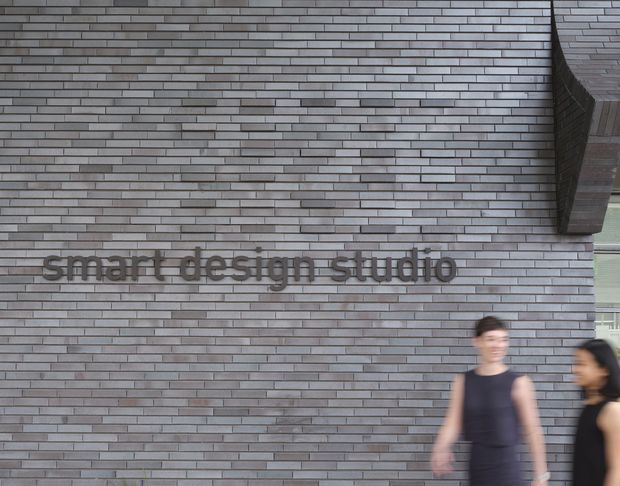 Exterior view of a contemporary commercial building facade clad in narrow horizontal grey brick slips, with raised dark lettering reading “smart design studio.” Two blurred pedestrians appear in the foreground, and a partial glazed entrance or window is visible at the right edge.