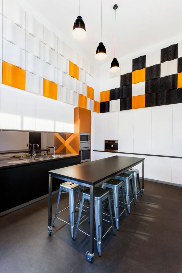 Bright, high-ceiling contemporary kitchen with extensive white flat-panel cabinetry, a long black island with a dark benchtop, stainless-steel stools, and integrated appliances. The room features bold black, white, and orange geometric acoustic or decorative wall panels, plus three suspended black pendant lights with white globes. Dark flooring and black cabinetry create strong contrast against the white surfaces.