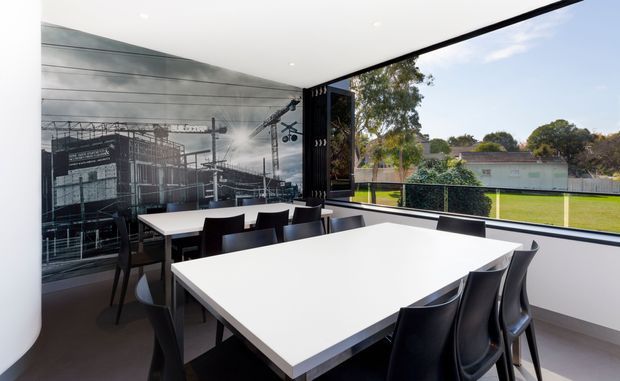 A bright contemporary dining room or meeting space with a large white rectangular table surrounded by black chairs. Floor-to-ceiling glazing opens to a green outdoor field, while a large monochrome construction-site mural forms a feature wall. The room has a clean, minimal fit-out with recessed ceiling lights and dark flooring.