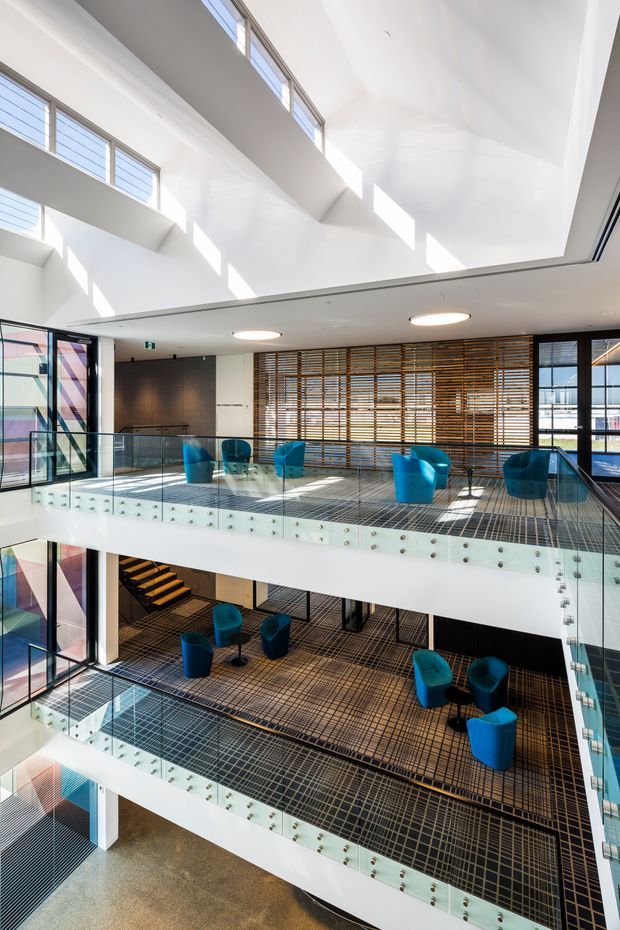 A bright, multi-level contemporary commercial interior with open atrium galleries, glass balustrades, exposed circulation stairs, and extensive clerestory glazing. Each floor contains informal lounge seating with vivid blue armchairs and small tables. Timber horizontal screening lines the upper-level wall, while patterned dark flooring and colourful glazed panels add visual interest. Strong daylight fills the tall void, creating an airy, modern communal space.