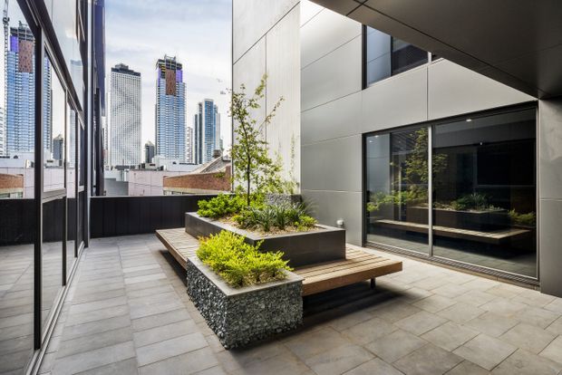 A contemporary urban rooftop terrace or courtyard with large-format grey paving, dark metal-framed glazing, and a minimalist built-in timber bench integrated around raised rectangular planters. The planters feature textured stone or pebble-clad sides and restrained green planting. Reflective glazing and tall city buildings are visible around the enclosed terrace, creating a polished high-rise setting.