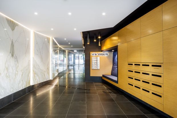 A polished commercial building lobby or apartment entry foyer with dark large-format tiled flooring, dramatic veined white feature wall panels, warm timber-toned built-in mailboxes and cabinetry, a dark angular ceiling feature, recessed downlights, pendant lighting, and glazed entrance doors at the far end. A seating niche and directory sign are integrated into the timber joinery. The space has a sleek, contemporary and refined character.