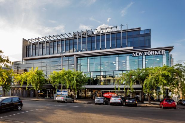 Street-facing photograph of a contemporary multi-storey commercial building with extensive blue-tinted glazing, a prominent rooftop pergola-like structural screen, and the 'NEW YORKER' sign on the facade. Ground-level retail and commercial frontage opens onto a tree-lined urban street with parked cars.
