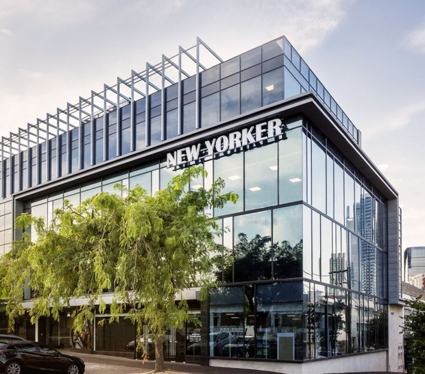 Exterior view of a multi-storey commercial building with a highly glazed curtain-wall facade, dark horizontal framing, projecting rooflines, and a distinctive exposed rooftop structural frame. The New Yorker sign is mounted above the ground-floor frontage, with trees and an urban high-rise context visible around the building.