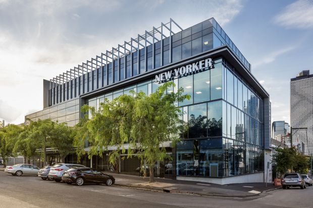 Street-level exterior view of a contemporary multi-storey commercial building with extensive blue-grey glass curtain walls, dark structural framing, and a prominent NEW YORKER retail sign. The building features a projecting glazed corner, rooftop linear steel framing, street-level retail frontage, surrounding urban streetscape, trees, and parked cars.
