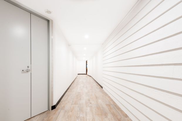 Bright, narrow contemporary hallway with pale timber-look flooring, white walls and ceiling, recessed downlights, a flush grey door on the left, and a distinctive white horizontally slatted feature wall on the right.