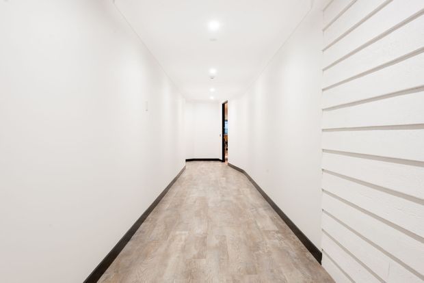 A long, narrow, bright white hallway with recessed ceiling downlights, light wood-look flooring, dark skirting boards, and a distinctive white horizontal slatted feature wall on the right. An open doorway is visible at the far end.
