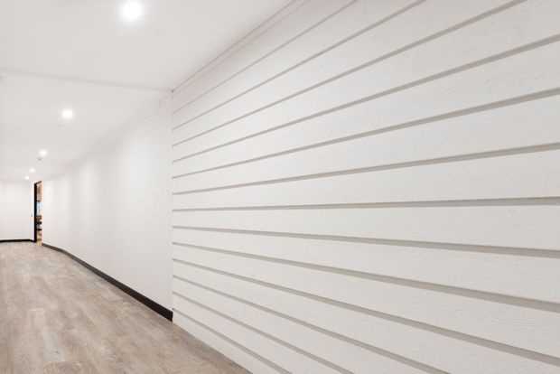 A bright, minimalist interior hallway with pale timber-look flooring, smooth white walls, recessed ceiling downlights, and a prominent white horizontally grooved feature wall. A dark skirting board runs along the base, with an open doorway visible at the far end.