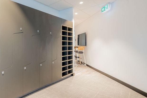 A clean, narrow interior corridor or staff area with a wall of dark grey built-in lockers and an adjacent open cubby shelving unit. At the far end is a small wall-mounted screen above a compact counter and stools. The space has pale carpet flooring, white walls and a suspended grid ceiling with recessed downlights and an illuminated emergency exit sign.