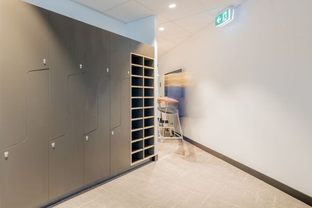 A clean commercial or institutional corridor with dark built-in locker cabinetry along the left wall, an open cubby shelving unit at the end, pale tiled flooring, white walls, recessed ceiling lights, and an illuminated emergency exit sign. A blurred person is moving through the hallway.
