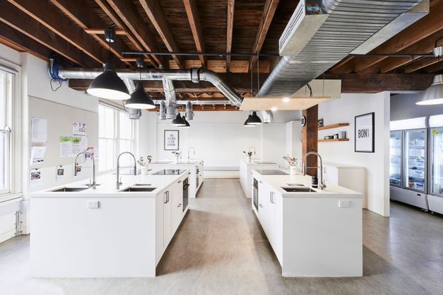 Bright open-plan commercial kitchen or culinary teaching space with multiple white island workbenches, sinks, taps and integrated appliances. The room has exposed dark timber ceiling joists, exposed metal ductwork, black pendant lights, white walls, polished concrete-toned flooring and large windows. Refrigerated display units are visible along the right wall.