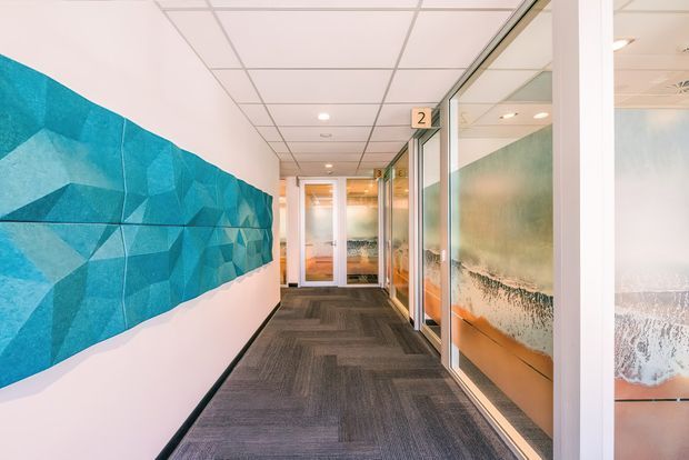 A bright contemporary commercial hallway with grey carpet tiles, white walls, a suspended acoustic tile ceiling, and glazed office partitions. A large faceted turquoise-blue feature wall mural runs along the left, while frosted and translucent glass panels with orange accents line the right side. Numbered room signs and recessed ceiling lights reinforce the office setting.
