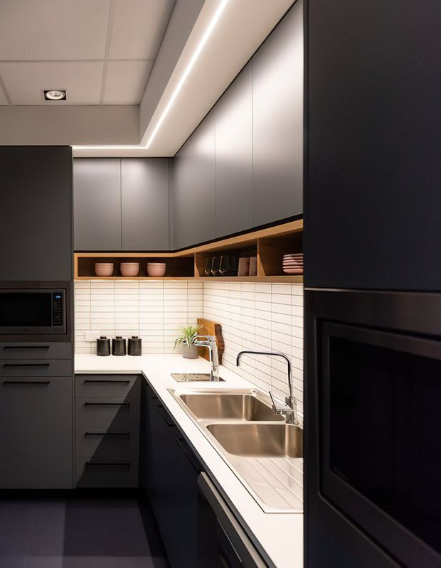 Compact contemporary galley kitchen with matte dark cabinetry, integrated appliances, a white horizontal-tile backsplash, warm timber open shelving, stainless-steel double sink, and continuous linear LED lighting beneath a tiled ceiling.