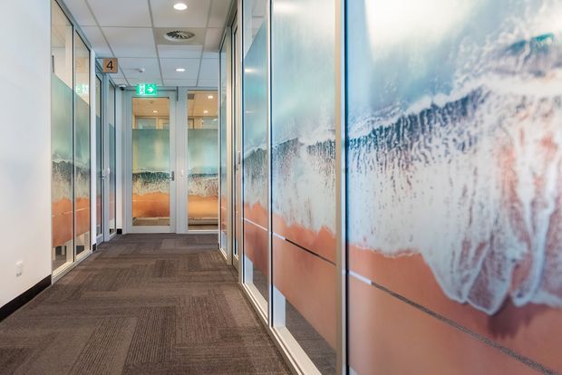 A modern commercial corridor with glass-partitioned offices on both sides, patterned carpet flooring, recessed ceiling lights, and a glass exit door at the end. The glazing features a large frosted abstract graphic in white, blue, and terracotta tones, creating a distinctive decorative feature wall effect.